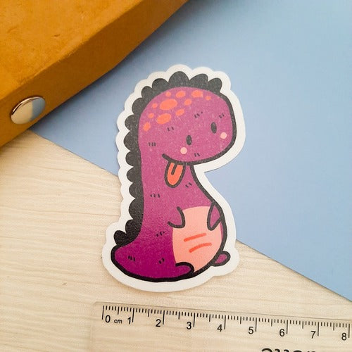 Kuvassa on tarra, joka on kieltä näyttävä violetti dinosaurus. Taustalla on ruskeaa, sinistä sekä valkoista. Kuvassa näkyy myös viivotin.