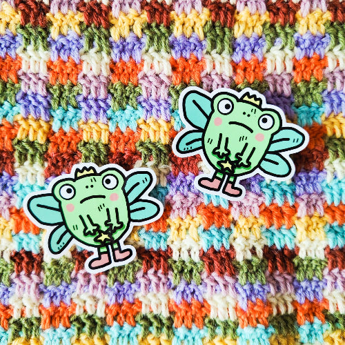 Kuvassa on sammakkotarra. Sammakolla on surullinen ilme ja silmät hieman kierossa. Sillä on myös keijun siivet, taikasauva, kruunu ja vaaleanpunaiset sukat.