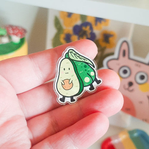 Kuvassa on käsi, joka pitelee pinssiä. Kuvassa on pinssi, jossa on kaksi toisissaan kiinni olevaa avokadoa. Vain toiselta näkyy etupuoli. Tämä avokado on tympäätyneen oloinen. Selkä katsojaan olevalla avokadolla sen sijaan näkyy pyllyvako.