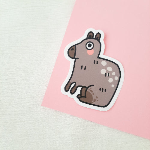 Kuvassa oleva tarra on harmaanruskea capybara. Sillä on suuret silmät ja vaaleanpunaiset posket.