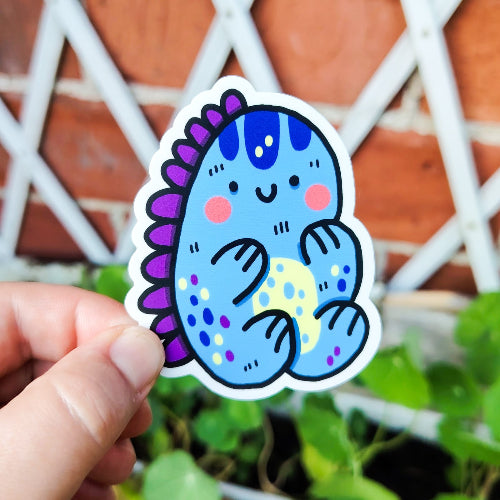 Kuvassa on sininen dinosaurustarra. Dinosauruksella on keltainen maha ja violetit selkävät. Dinosaurus on hieman pyöreä ja sillä on iloinen ilme.