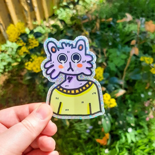 Kuvassa oleva tarra on violetti kissa, jonka silmistä valuu kyyneleitä. Kissalla on suuret silmät ja keltainen paita päällä. Tarralla on glitteriset reunukset.