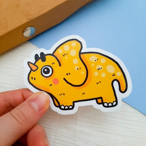 Kuvassa on henkilön käsi pitelemässä keltaista dinosaurustarraa. Taustalla on ruskea penaali, sininen pala kartonkia ja valkoinen kangas.