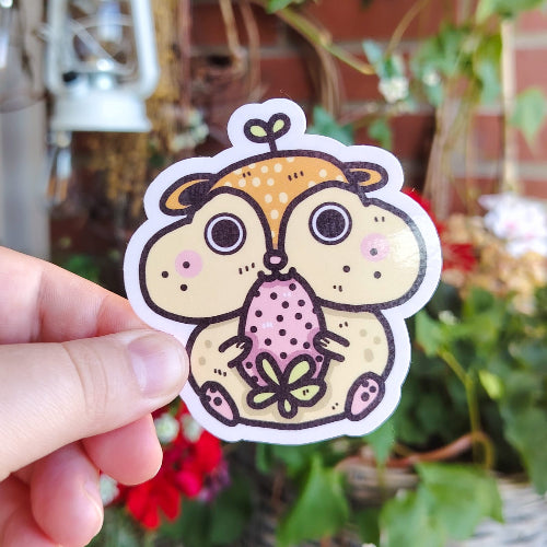 Kuvassa on vaaleanbeige hamsteritarra. Hamsterilla on jättimäiset posket, ja se koittaa mutustella oamn päänsä kokoista mansikkaa.