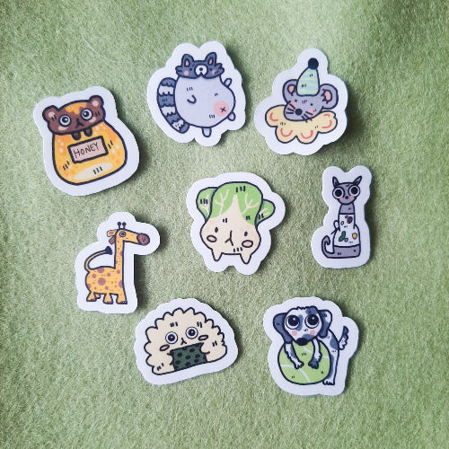 Kuvassa on kahdeksan pientä ja hauskaa eläin- ja ruokatarraa vihreän pohjan päällä.