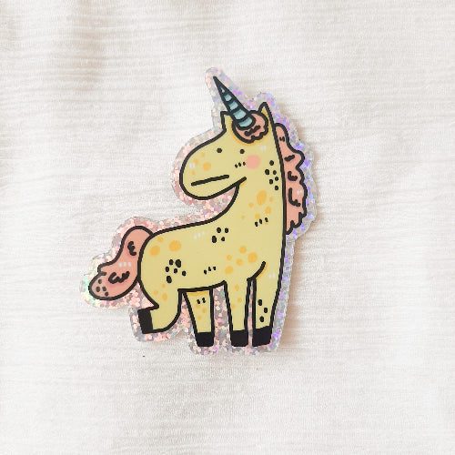 Kuvassa oleva tarra on hauska yksisarvinen, jolla on hieman tympeä ilme. Yksisarvisella on sininen sarvi ja vaaleanpunaiset harjas ja häntä. Yksisarvisen kaviot ovat mustat ja se nostaa yhtä jalkaansa ylöspäin elegantisti.