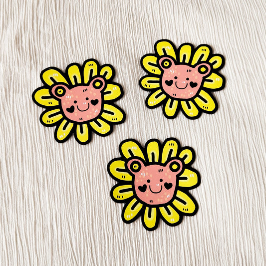 Kuvassa on hauska karhumagneetti. Magneetissa on vaaleanpunainen karhun pää, jonka ympärillä on keltaisia kukan terälehtiä.