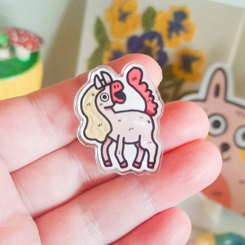 Kuvassa on käsi pitelemässä pinssiä. Pinssi on vaaleanpunainen hevonen, jolla on vaaleanruskea harjas ja pinkki häntä.