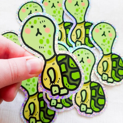 Kuvassa oleva tarra on vihreä kilpikonna. Tarran reunus on glitterinen. Kilpikonnalla on hieman surullinen ilme, mutta se näyttää silti hauskalta.