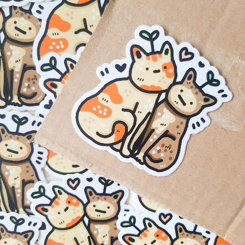 Kuvassa on kaksi kissaa. Toinen kissa on vaalea, jossa on oransseja laikkuja ja toinen on siamilainen kissa. Kissat kiehnäävät toisiaan vastaan, ja niillä on totinen ilme. Molempien kissojen päästä kasvaa kasvi.