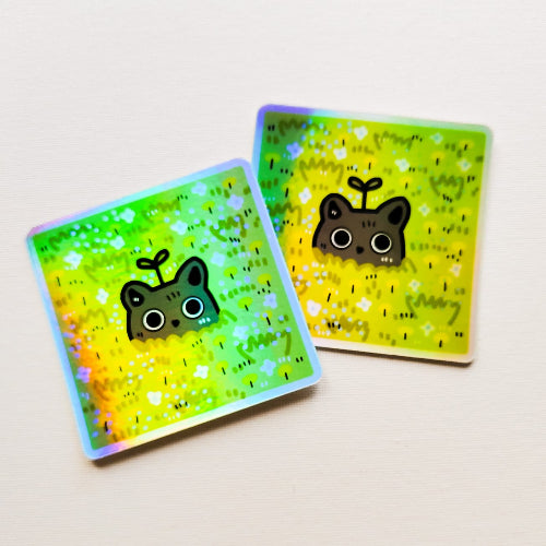 Kuvassa on holograafinen neliön muotoinen kissatarra. Musta kissa kurkkii kukkapellosta ja sen päästä kasvaa hauska pieni kasvi.