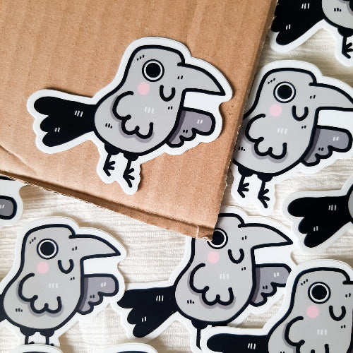 Kuvassa oleva vedenkestävä tarra on harmaa korppi, joka hymyilee iloisesti. Korpilla on suuret silmät ja pinkit, kirkkaat posket. Hauskan linnun peräsulat ovat oikein suuret ja komeat.