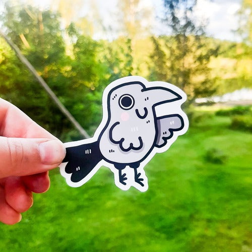 Kuvassa oleva vedenkestävä tarra on harmaa korppi, joka hymyilee iloisesti. Korpilla on suuret silmät ja pinkit, kirkkaat posket. Hauskan linnun peräsulat ovat oikein suuret ja komeat.