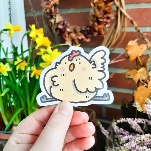 Kuvassa on hauska tarra. Tarra on beige kana, joka venyttelee. Sillä on tyhjä ilme ja tuuheat höyhenet.