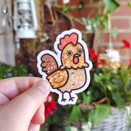 Kuvassa on kanatarra. Kana on ruskea, ja sillä on punaiset heltat ja keltainen nokka. Kanalla on myös suuret silmät ja hauskat jalat.