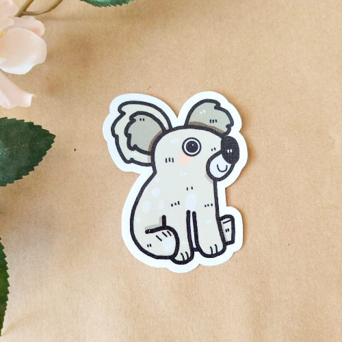 Kuvassa on vaaleanharmaa koalatarra. Koala hymyilee ja sillä on suuret silmät.