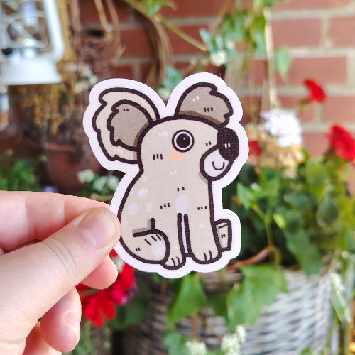 Kuvassa on vaaleanharmaa koalatarra. Koala hymyilee ja sillä on suuret silmät.