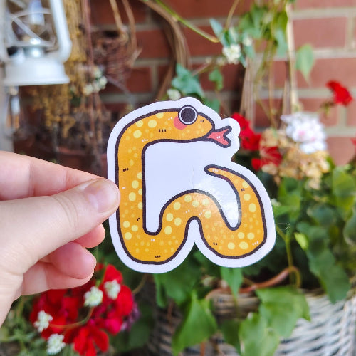 Kuvassa on oranssi käärmetarra. Käärme on on pitkä ja kiemurainen, ja siinä on keltaisia laikkuja. Käärmeellä on suuret silmät, vaaleanpunaiset posket ja kirkkaanpunainen pitkä kieli.