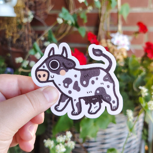 Kuvassa on hauska lehmätarra. Lehmä on valkoinen, ja siinä on perinteisiä mustia laikkuja. Lehmä hymyilee ja sillä on suuret silmät.