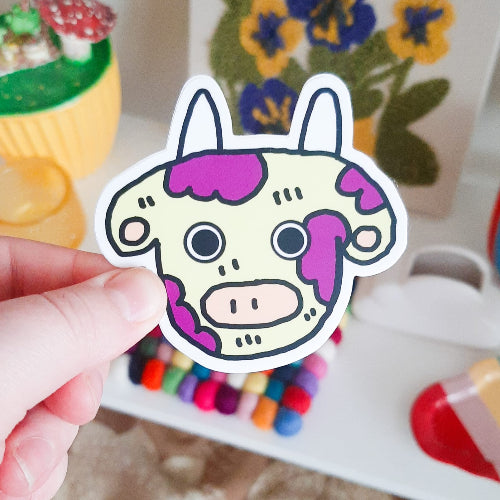 Kuvassa oleva tarra on lehmä, jonka pohjaväri on vaalean keltainen ja sillä on suuret, liilat laikut. Lehmällä on suuret silmät ja mahtavat, valkoiset sarvet.