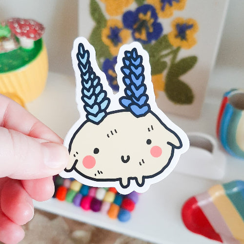 Kuvassa oleva tarra on beige olento, jonka päästä kasvaa kaksi sinistä kasvia. Mönttimäinen olento hymyilee iloisesti ja sillä on kirkkaasti loistavat pinkit posket.