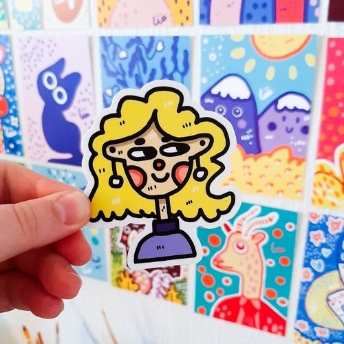 Kuvassa oleva tarra on hauskan näköinen nuori nainen. Hänellä on valkoiset roikkuvat korvakorut ja sininen paita. Naisen hiukset ovat keltaiset ja hän hymyilee.