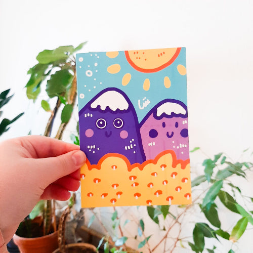 Kuvassa on postikortti, jossa on kaksi liilaa vuorta. Taustalla paistaa aurinko ja kuvituksen etualalla on keltainen puska.