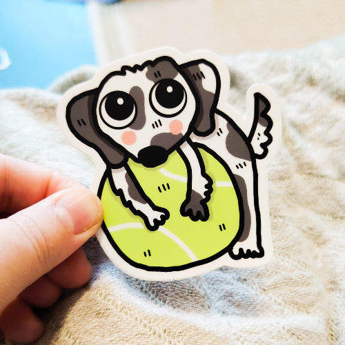 Kuvassa oleva tarra on dalmatiankoiran pentu halaamssa isoa, vihreää tennispalloa. Dalmatialainen tuijottaa katsojaa suurilla sympaattisilla silmillään.