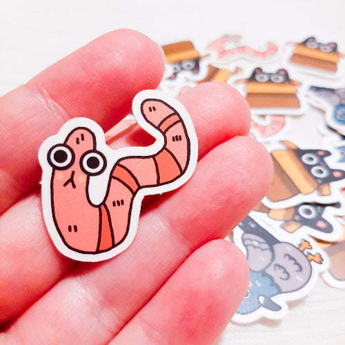 Kuvassa on pieni 3 cm kokoinen tarra. Tarra on hauska vaaleanpunainen mato, jolla on suuret silmät.