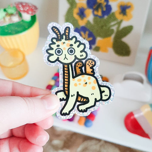 Kuvassa oleva tarra on keltainen lohikäärme, jolla on ihmettelevä ilme.
Lohikäärmeessä on oransseja kuviointeja, kuten siivet, sarvet ja maha. Lohikäärmeellä on isot silmät ja hieman harmistunut ilme. Tarran reunus on glitterinen.