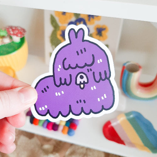 Kuvassa oleva tarra on liila koira. Koiran pitkät karvat peittävät sen silmät ja naaman. Sen suusta tulee ulos pieni vaaleanpunainen kieli.