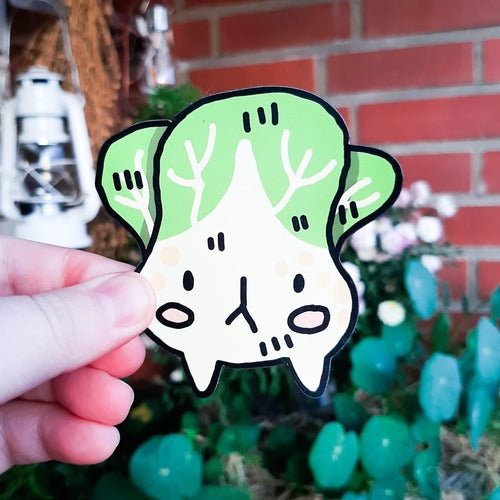 Kuvassa henkilön käsi pitelee magneettia. Magneetti on surullisen näköinen bok choy eli suomeksi pinaattikiinankaali. Tällä hauskalla vihannesmagneetilla on komeat, vaaleanpunaiset magneetit ja kaksi pientä jalkaa.
