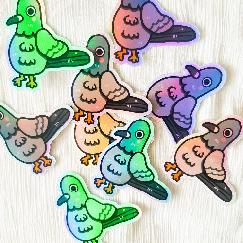 Kuvassa on pulutarra. Tarra on holograafinen, ja se loistaa sateenkaaren väreissä, kun siihen osuu aurinko. Pulu on harmaa, ja sillä on oikein komeat sulat, isot silmät ja vaaleanpunaiset posket.