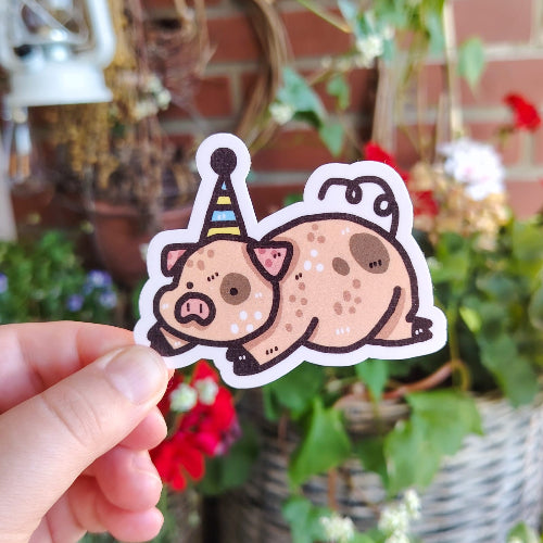 Kuvassa on possutarra. Possu on vaaleanpunainen, ja siinä on pari ruskeaa läikkää. Possulla on kippurahäntä, se makaa mahallaan ja sillä on juhlahattu päässä.