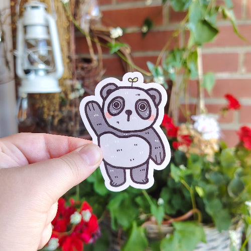 Kuvassa on hauska pandatarra. Panda on harmaavalkoinen, ja se nostaa toista tassuaan tervehdykseksi. Panda seisoo ja sillä on suloiset kiiluvat silmät. Sen päästä kasvaa pieni kasvi.