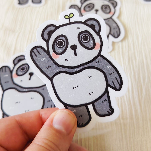 Kuvassa on hauska pandatarra. Panda on harmaavalkoinen, ja se nostaa toista tassuaan tervehdykseksi. Panda seisoo ja sillä on suloiset kiiluvat silmät. Sen päästä kasvaa pieni kasvi.