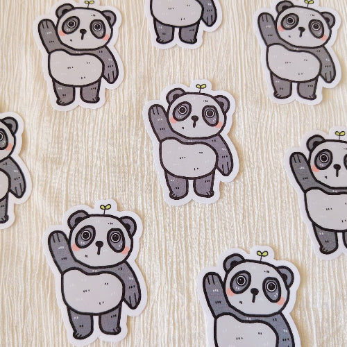 Kuvassa on hauska pandatarra. Panda on harmaavalkoinen, ja se nostaa toista tassuaan tervehdykseksi. Panda seisoo ja sillä on suloiset kiiluvat silmät. Sen päästä kasvaa pieni kasvi.