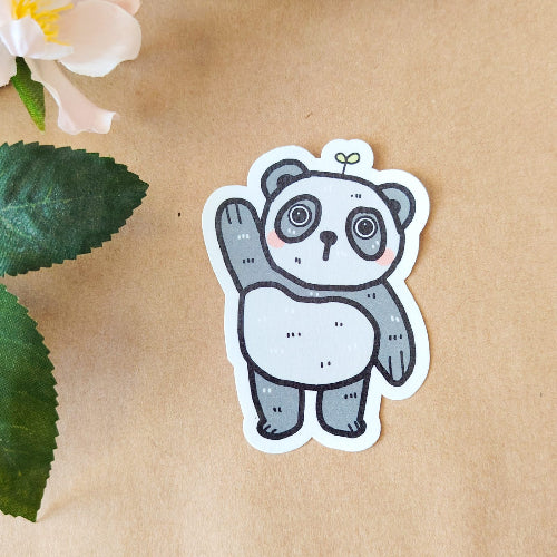 Kuvassa on hauska pandatarra. Panda on harmaavalkoinen, ja se nostaa toista tassuaan tervehdykseksi. Panda seisoo ja sillä on suloiset kiiluvat silmät. Sen päästä kasvaa pieni kasvi.