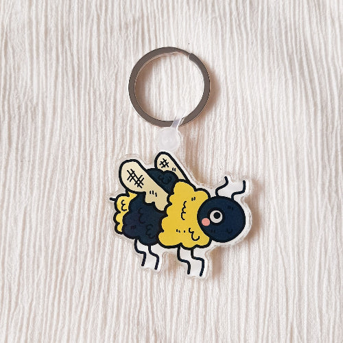 Valkoisella kankaalla on akryyinen avaimenperä, joka on mehiläinen. Tämä hauska pörriäinen on oikein karvainen ja hymyilee iloisesti.