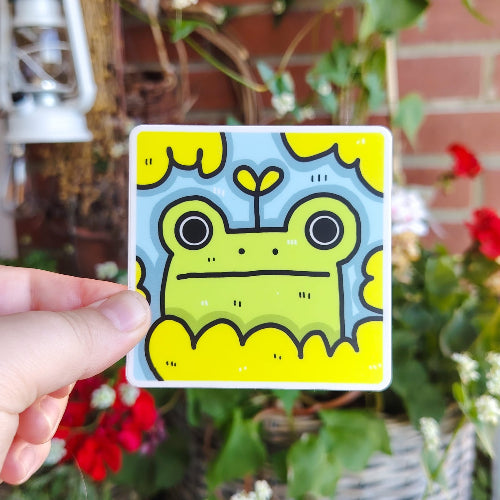 Neliön muotoinen sammakko, jossa on vihreä sammakko. Sammakolla on suuret silmät, totinen ilme ja sen päästä kasvaa pieni kasvi.