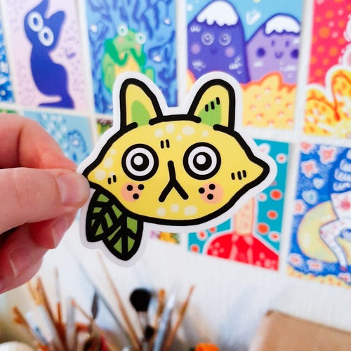 Kuvassa on tarra, joka on keltainen sitruunan muotoinen kissan pää. Kissalla on mietteliäs ilme ja suuret korvat. Tarrassa on myös kaksi vaaleankeltaista lehteä.