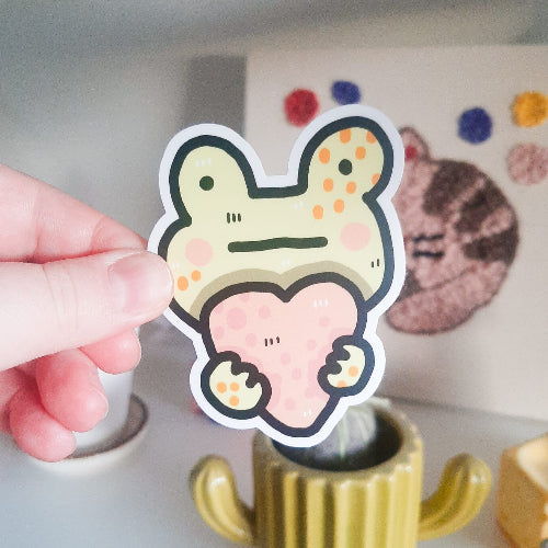 Kuvassa oleva tarra on hauskan näköinen sammakko halaamassa vaaleanpunaista sydäntä. Sammakolla on vähän tympääntynyt ilme. Sammakko on keltainen ja sydän vaaleanpunainen.