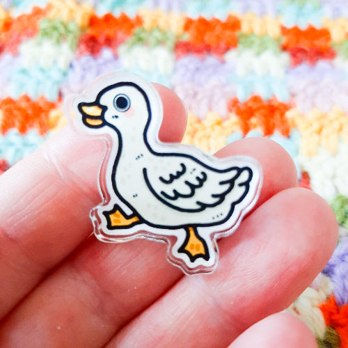 Kuvassa on yksi hauskan näköinen ankkapinssi. Ankalla on oranssi nokka ja räpylät, ja se näyttää vaappuvan hauskasti eteenpäin. Käsi pitelee pinssiä ja kuvan taustalla on värikäs virkkaus.