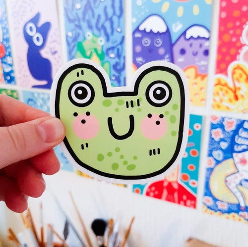 Kuvassa on tarra, joka on vihreä sammakkko. Sammakko hymyilee ja sillä on kirkkaat pinkit posket. Sen silmät ovat suuret.