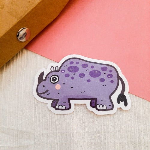 Kuvassa on tarra, jossa on iloinen liila sarvikuono. Taustalla on pinkkiä.