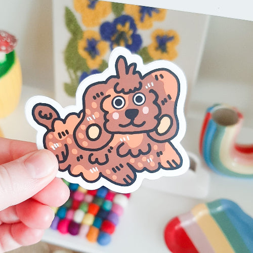 Kuvassa on käsi, joka pitelee koiratarraa. Tarra on iloinen spanieli, jonka turkki on oikein karvainen ja kikkara.