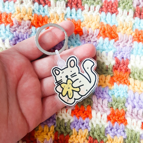 Käsi pitelemässä vaaleaa kissa-avaimenperää. Taustalla on värikäs ja pirteä kangas. Beige kissa istuu pyllyllään, halaten keltaista, hymyilevää tähteä. Kissalla on komeat viiksikarvat, vaaleanpunaiset korvat ja oikein iso häntä.