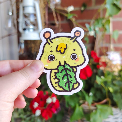 Kuvassa on hauska limenvihreä toukkatarra. Toukka mutustelee kirkkanvihreää lehteä ja hymyilee.