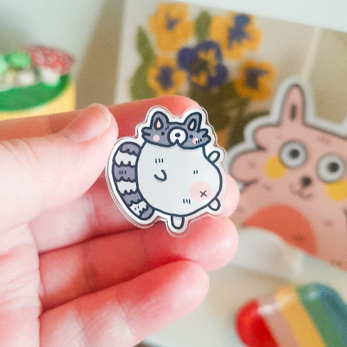 Pinssi, jossa on pyöreä pesukarhu. Sillä on vaaleanpunainen maha sekä posket. Pesukarhulla on myös oikein tuuhea häntä.