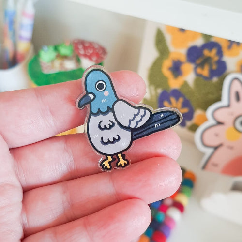 Harmaasävyinen pulupinssi. Pululla on pitkät peräsulat jsa oranssit jalat. Hänelllä on hieman hämmentyneen oloinen ilme. 